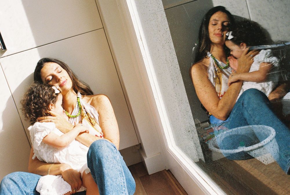 Woman sitting on floor cradling a child, with mirror reflection showing her holding a baby near a blue basin.