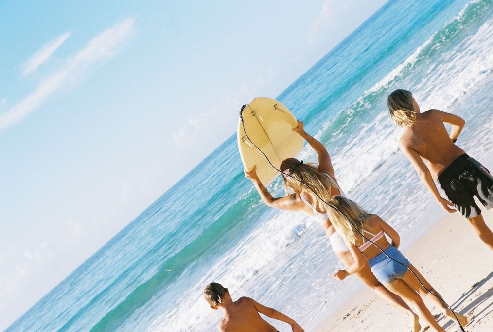 Group of friends at the beach with a yellow surfboard heading toward ocean waves on a sunny day.