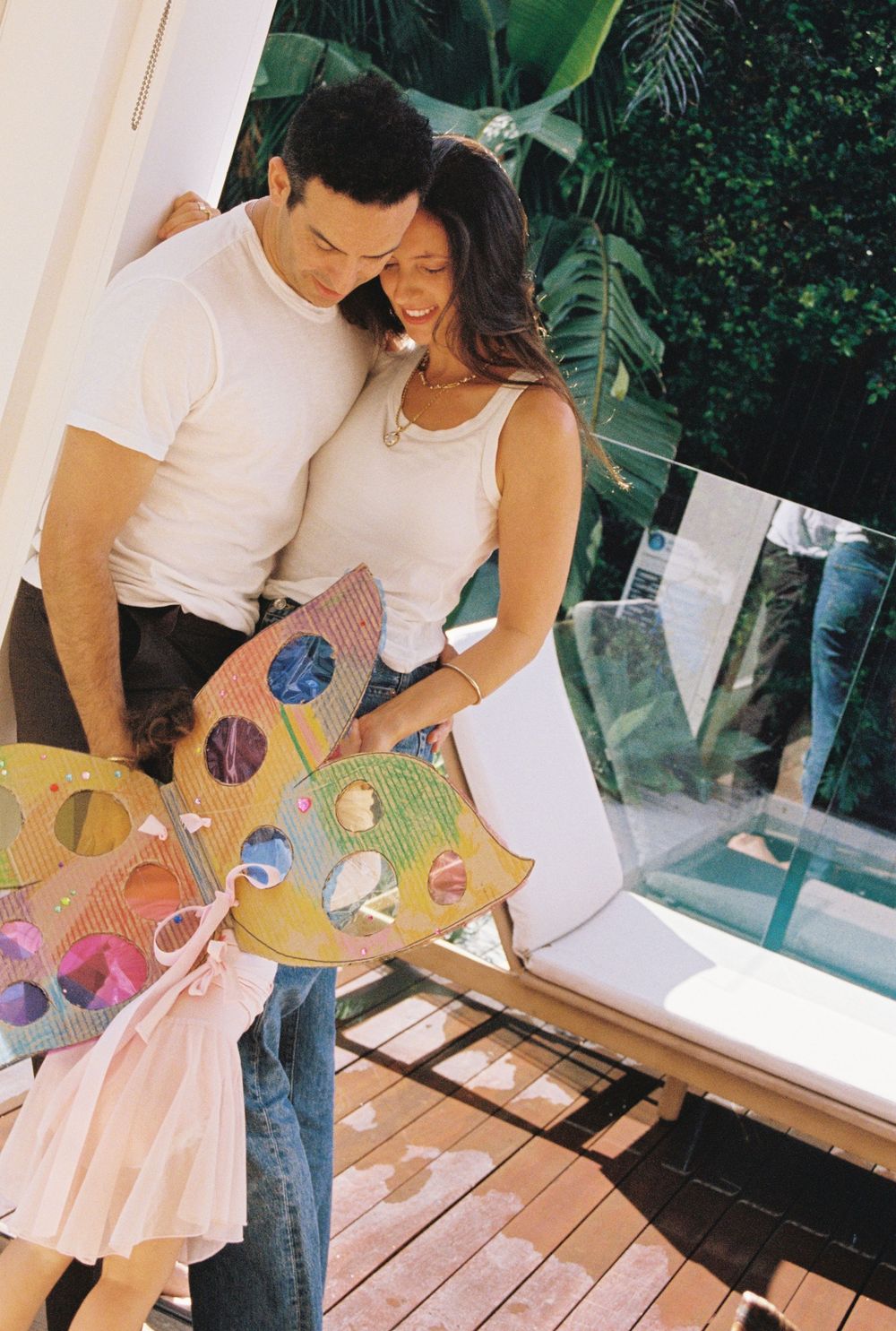 A couple smiles at a child holding a colorful paint palette on a sunny outdoor deck surrounded by tropical greenery.