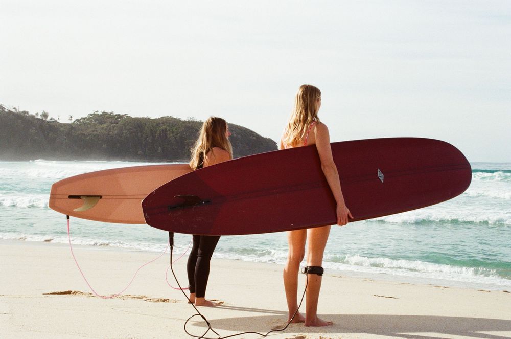 A surfer walks along a sandy beach carrying a red longboard surfboard with ocean waves in the background.