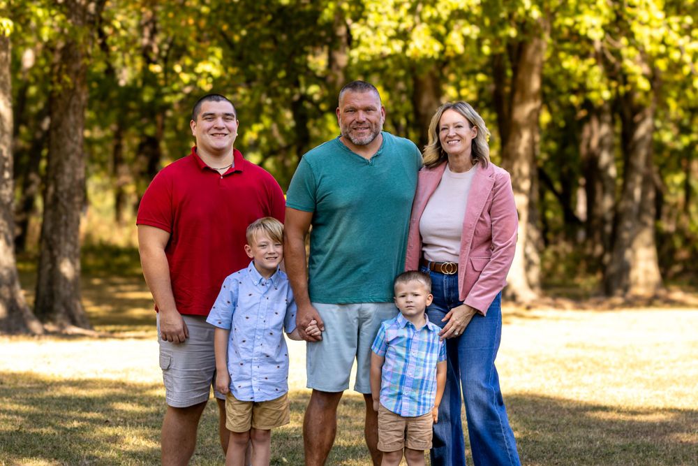 Family of five poses together for outdoor portrait in casual attire against wooded background.