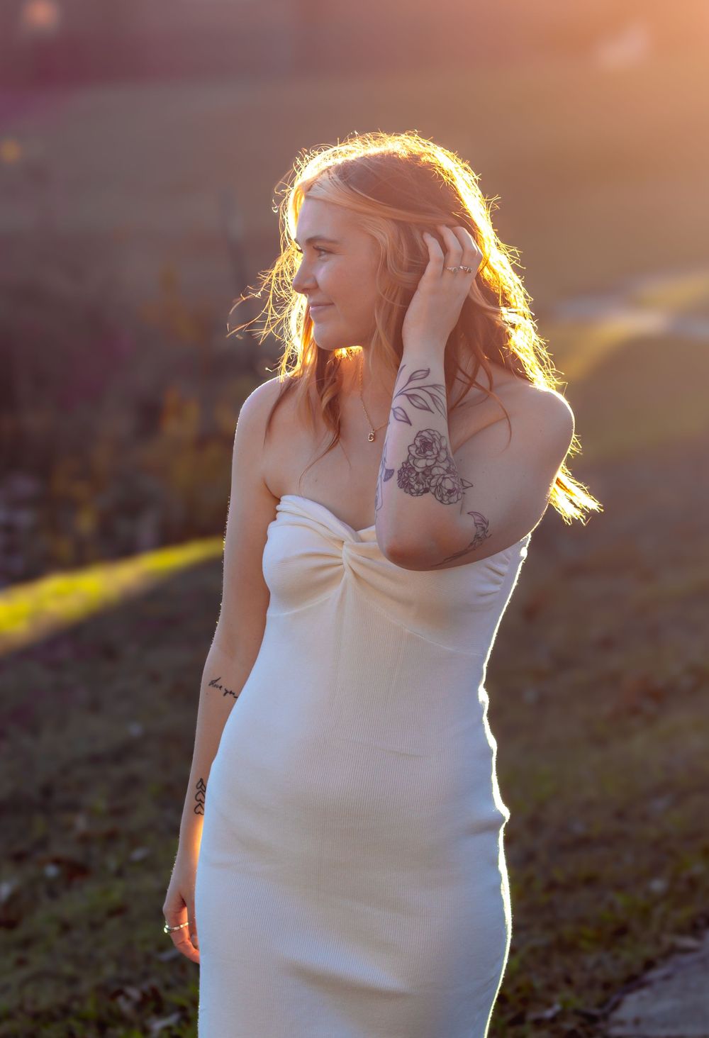 A pregnant woman in a white dress stands in golden sunlight during sunset, showing her silhouette and tattoos.