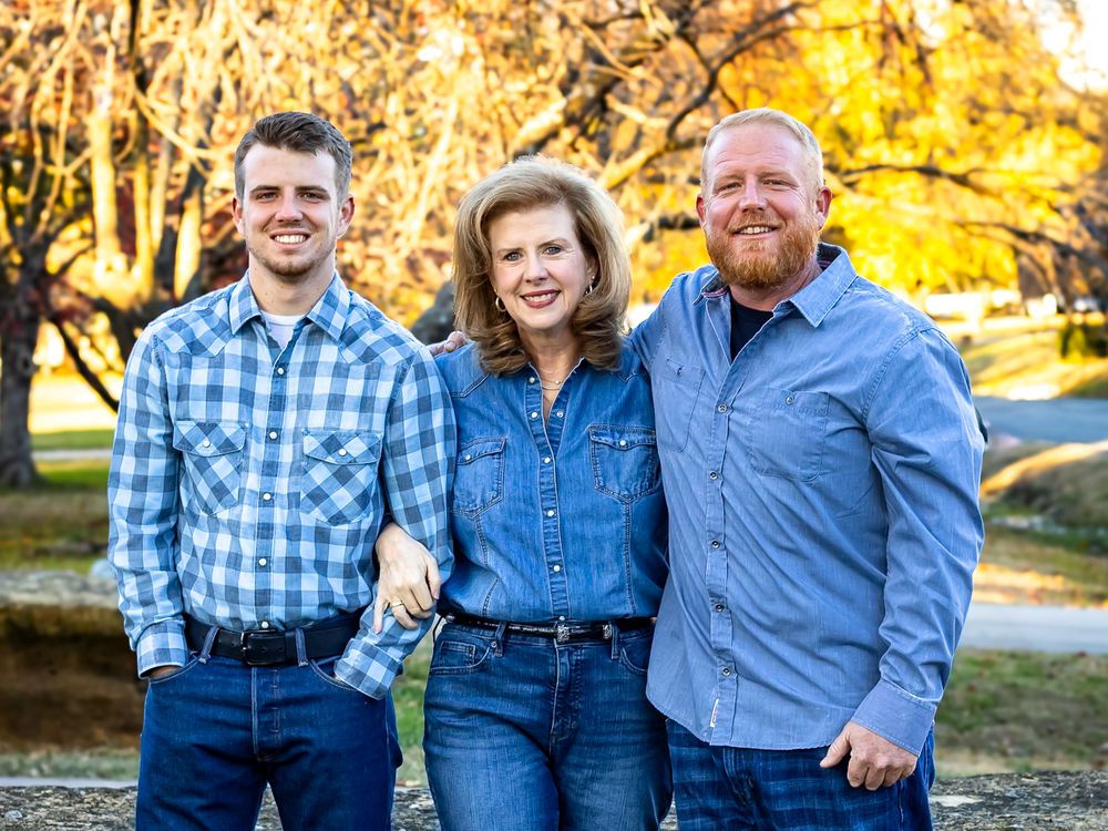 Three people in denim and blue shirts pose together outdoors during golden autumn afternoon light.