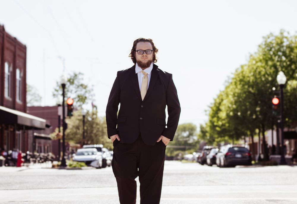 Person wearing a black suit and glasses stands confidently on a city street with storefronts and trees in the background.