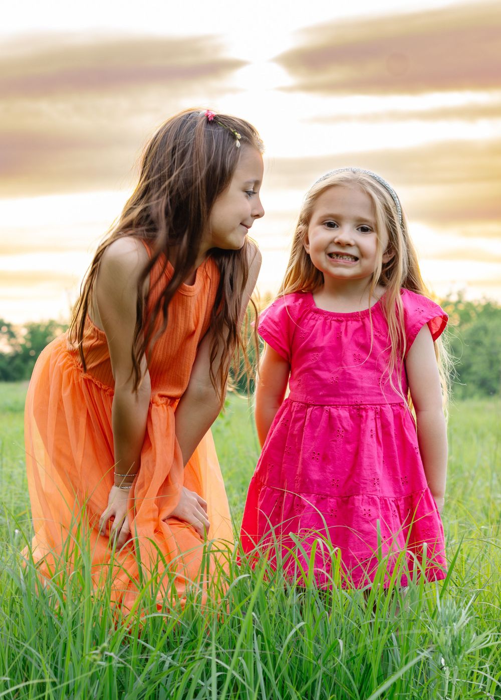 Two children in bright dresses playing together in tall grass during sunset.