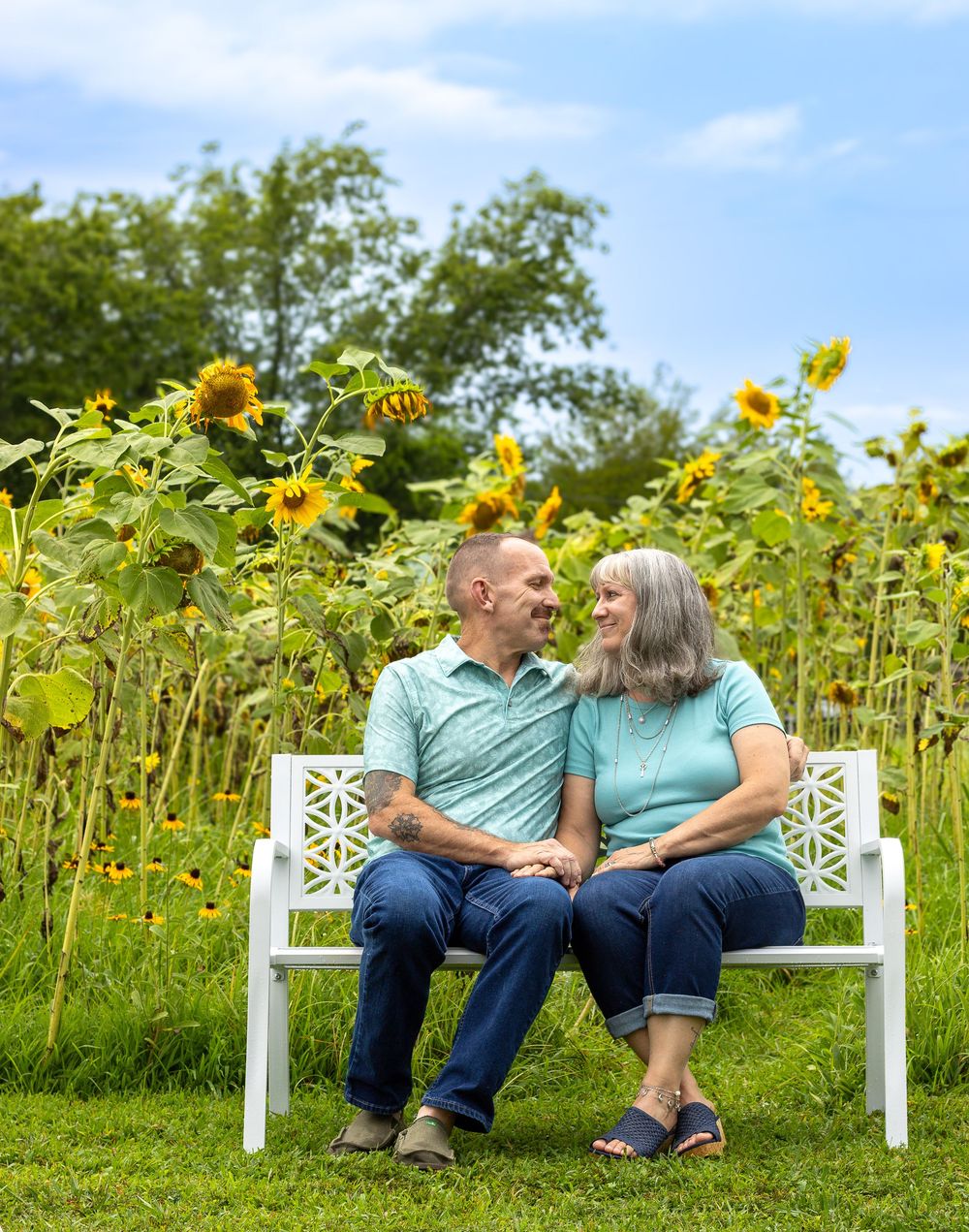 A couple sits lovingly on a white garden bench surrounded by tall blooming sunflowers on a sunny summer day.