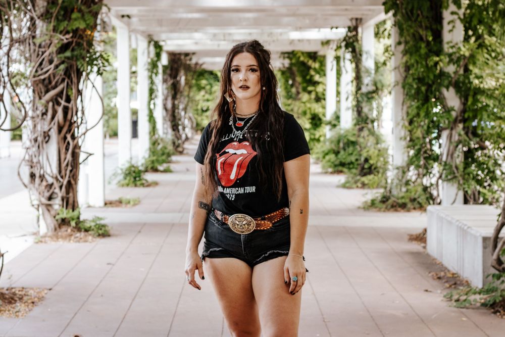 Someone wearing a Rolling Stones t-shirt and denim shorts poses in an outdoor corridor with white pillars and climbing vines.