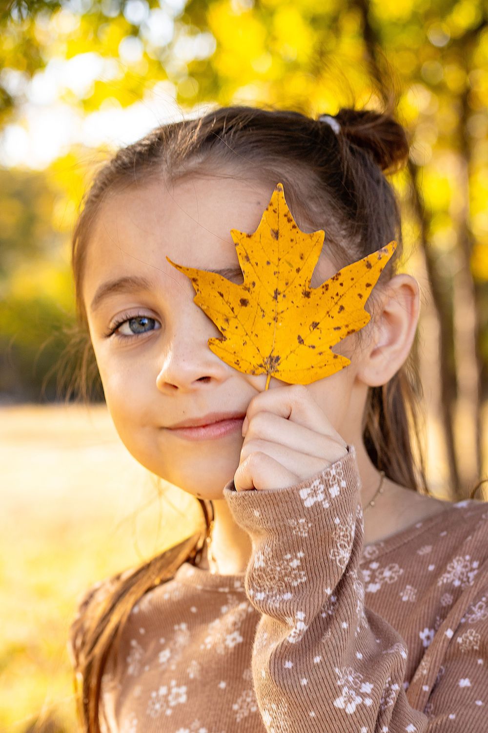 A bright yellow autumn maple leaf is held up against a sunny fall background with warm golden bokeh.