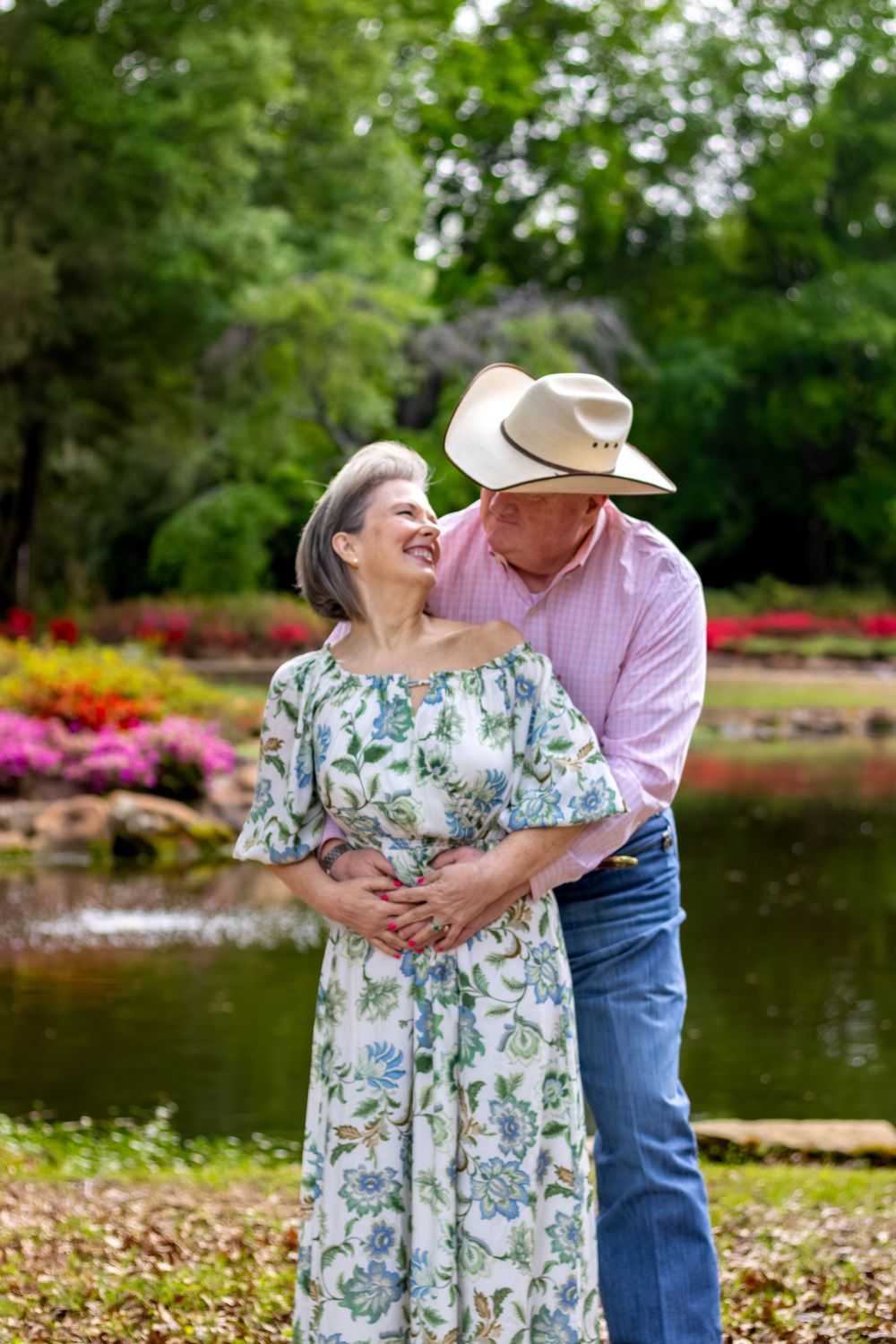 A smiling older couple embraces in a lush garden, man in cowboy hat and pink shirt, woman in floral dress near a pond.