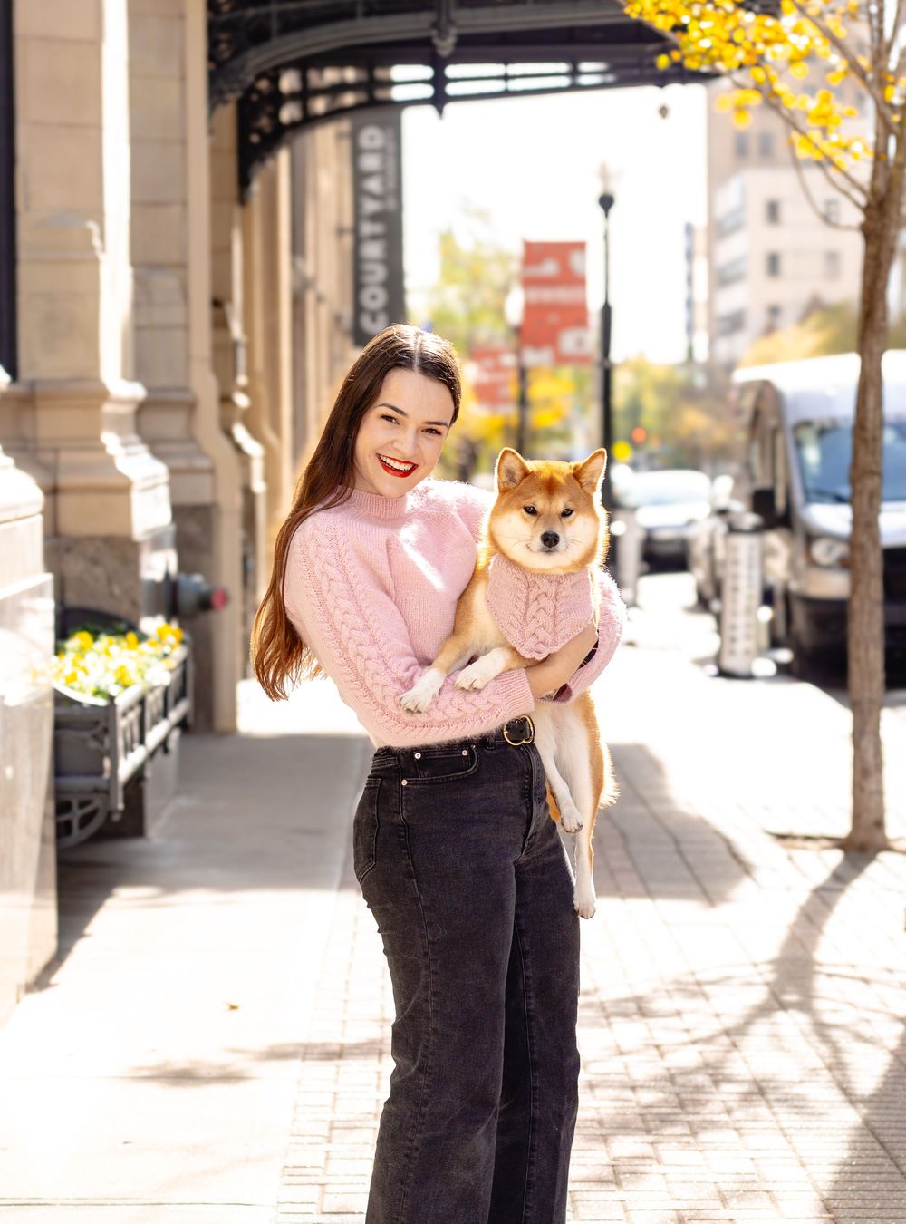 A person in a pink sweater holds a happy Shiba Inu dog while standing on a city sidewalk with autumn trees in the background.