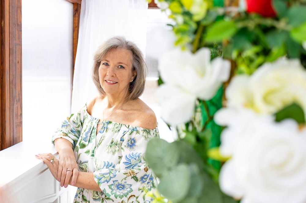 A person in a floral off-shoulder dress poses by a window with white flowers in the background.