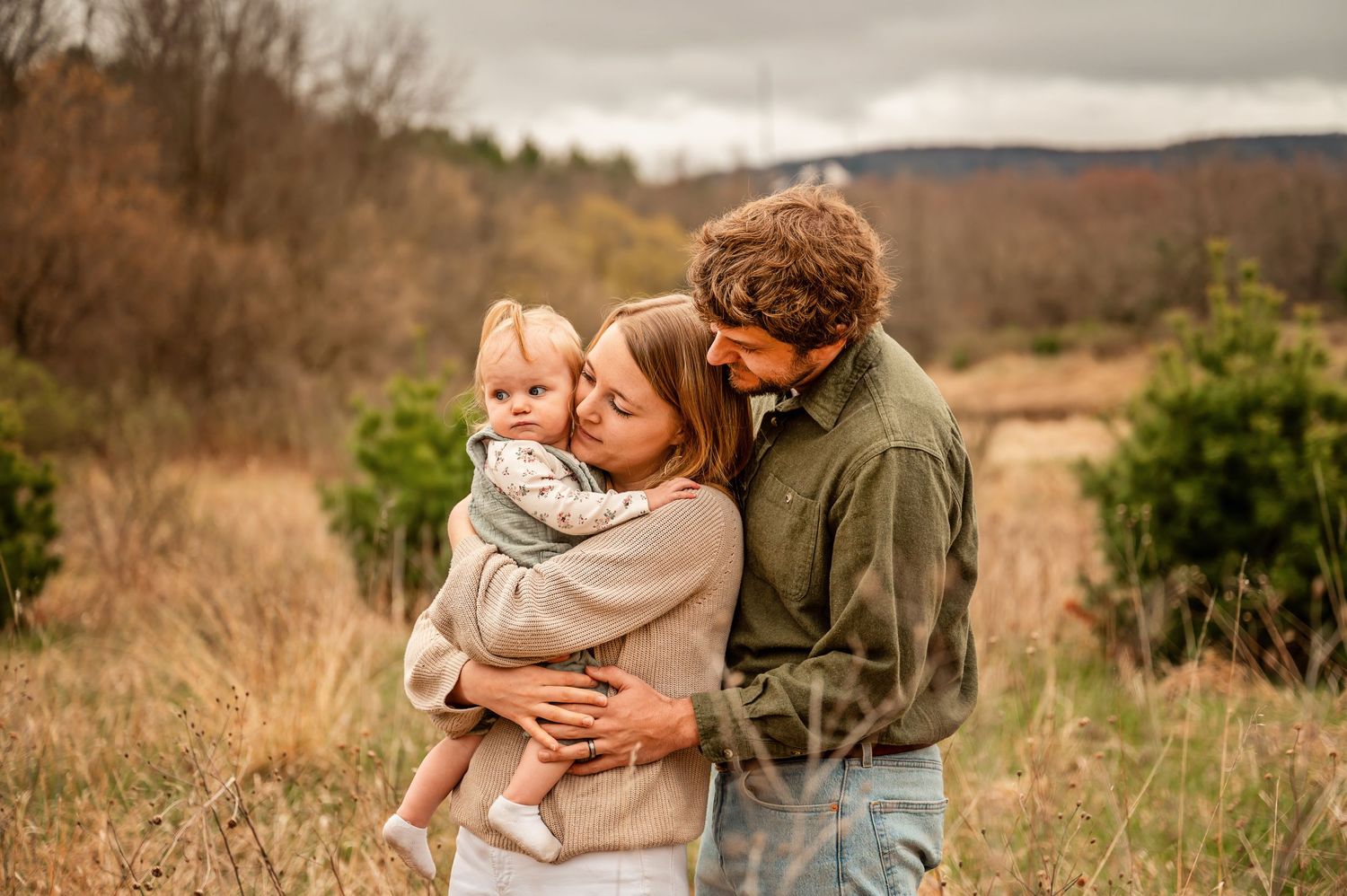 A tender family moment captured outdoors in a rustic field with mountains visible in the background on an autumn day.