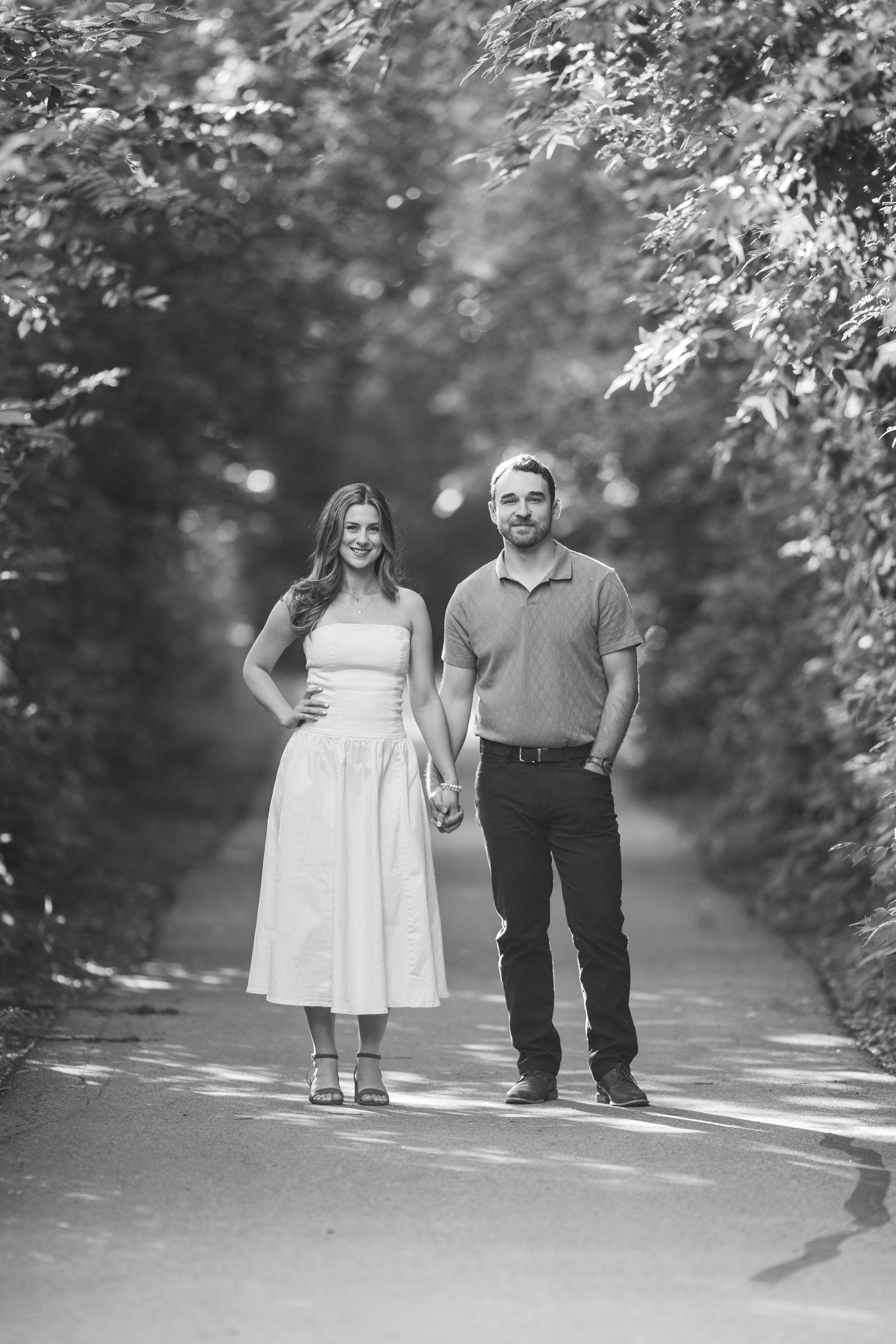 A couple walks hand in hand down a tree-lined path in black and white photography.