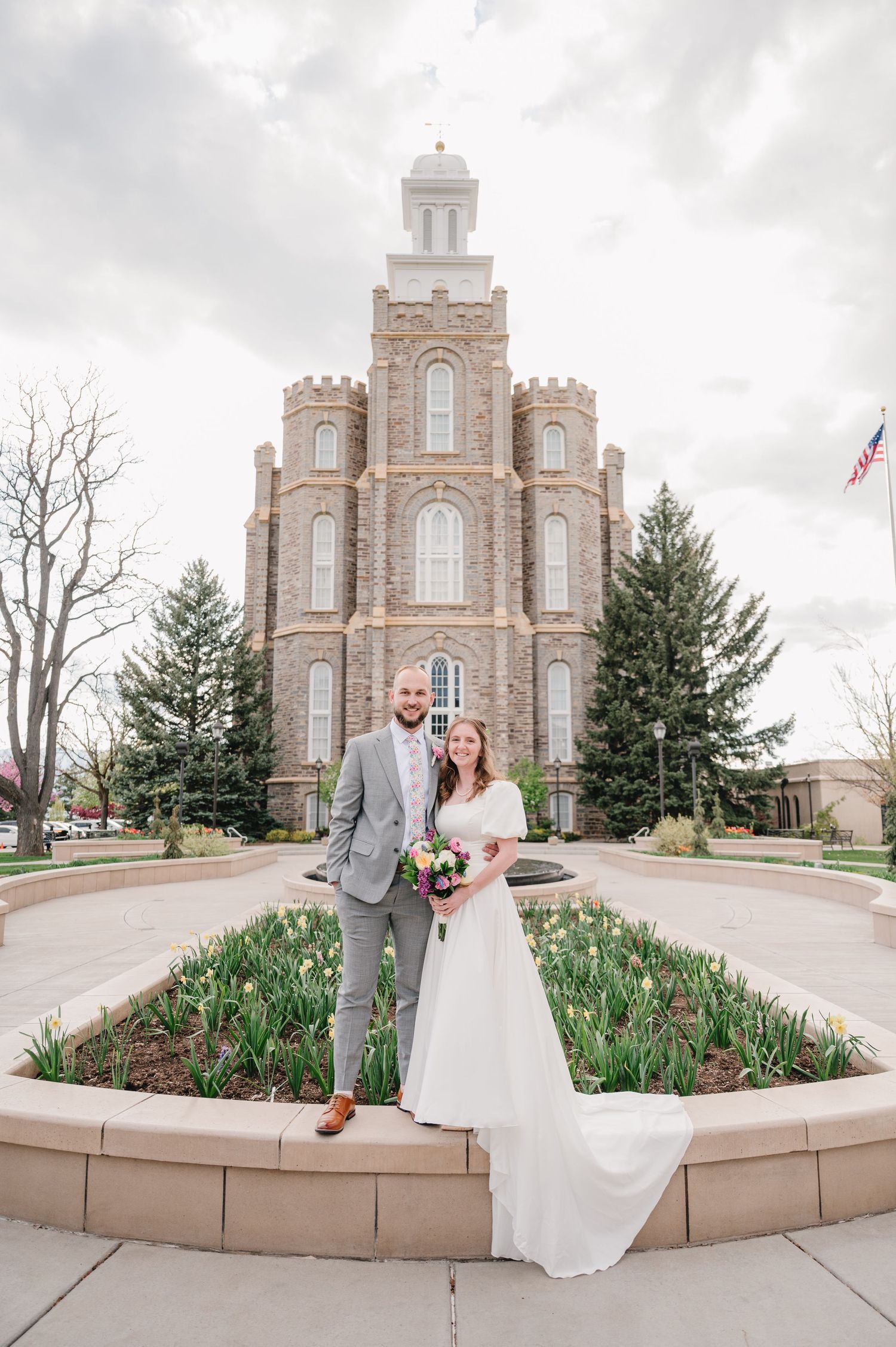 A wedding photo in front of the Logan Utah Temple with tall spires and Gothic architecture against a cloudy sky.