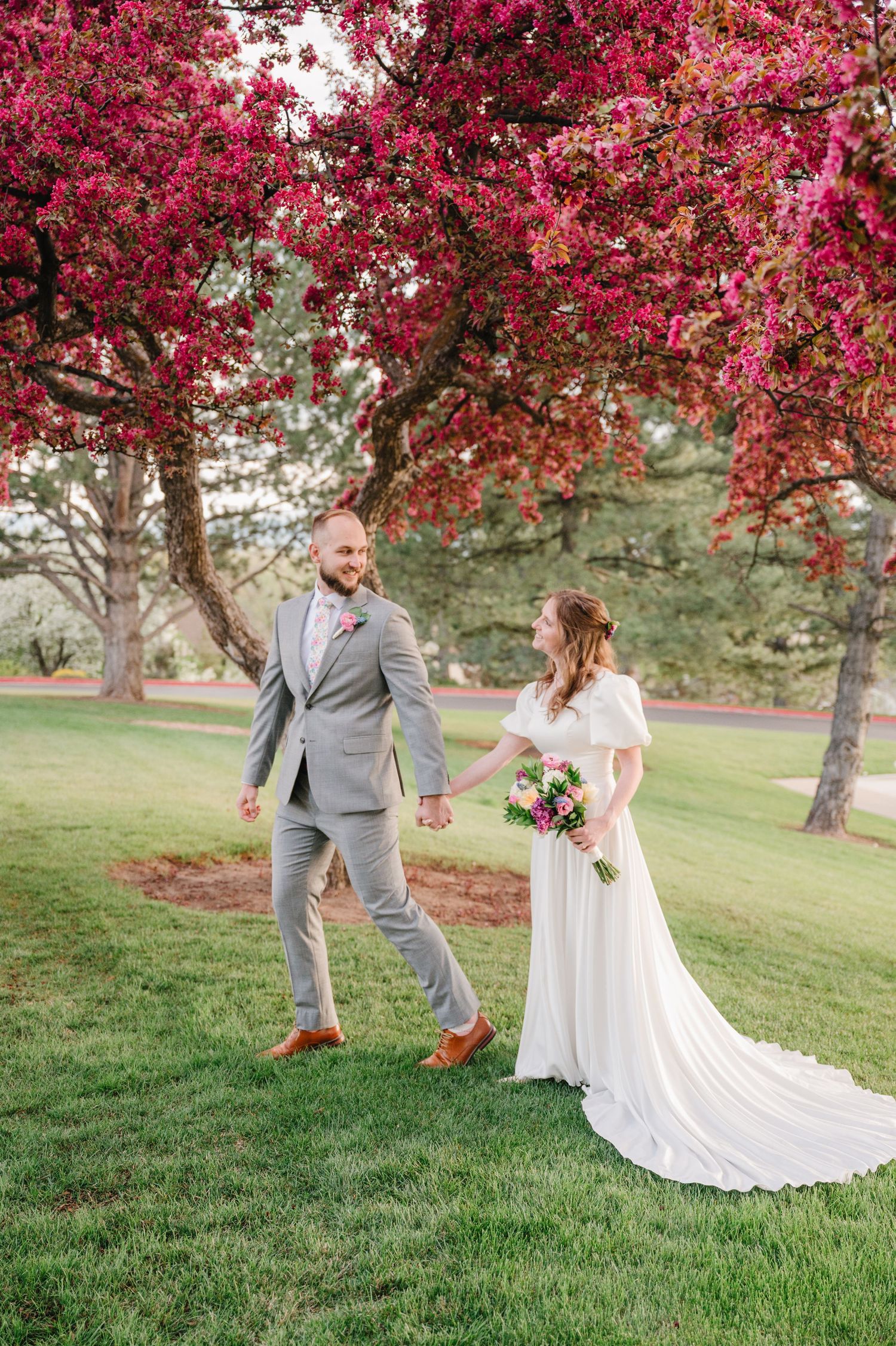 A couple in wedding attire walks joyfully under vibrant pink flowering crabapple trees on a grassy lawn.