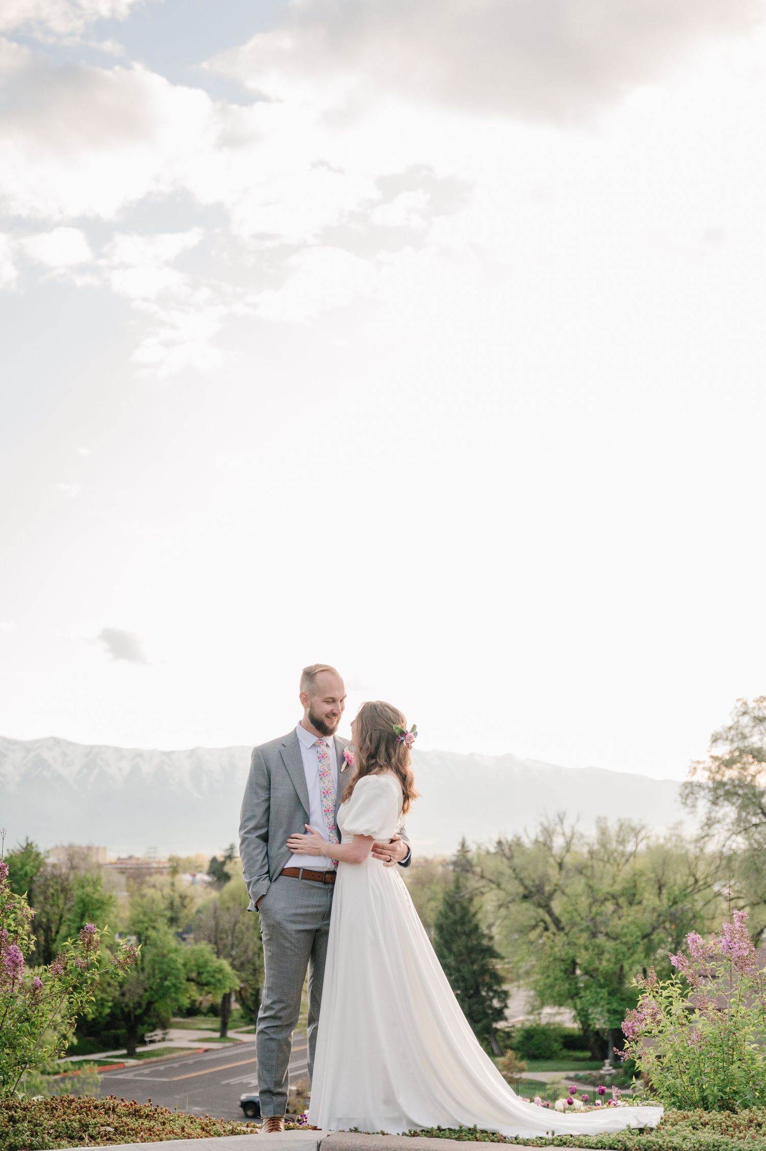 A couple embraces in a garden with mountain views on a cloudy day in elegant wedding attire.
