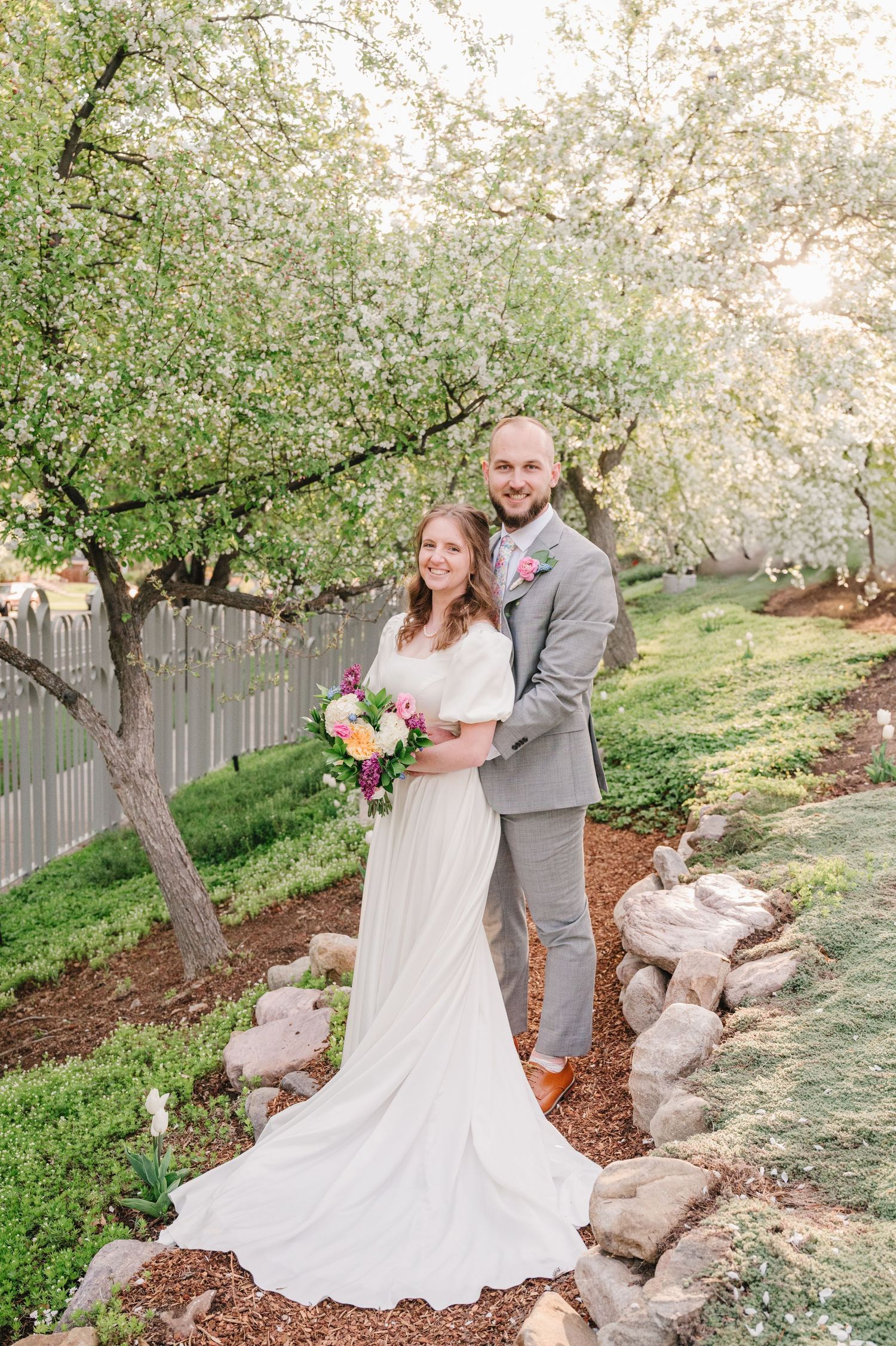 A couple poses together under blooming spring trees in an outdoor garden with white fence and stone landscaping.