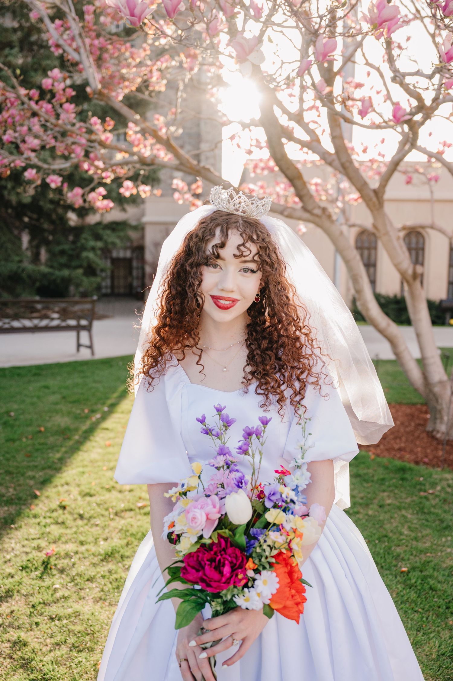 Beautiful bride in white dress holds vibrant spring bouquet under blooming magnolia trees in garden setting.