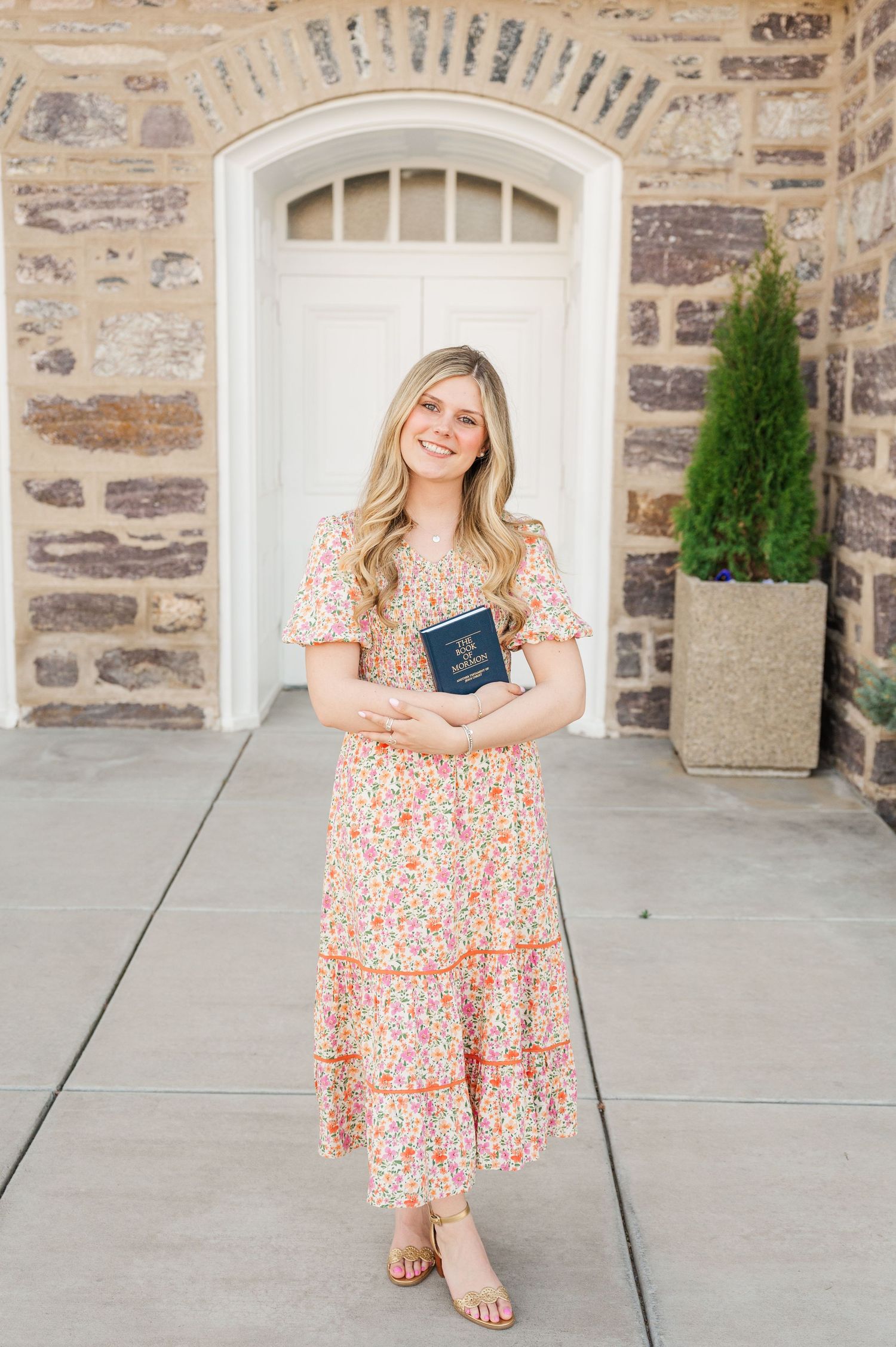 Woman in floral dress holds navy book while standing by stone wall and white doorway.