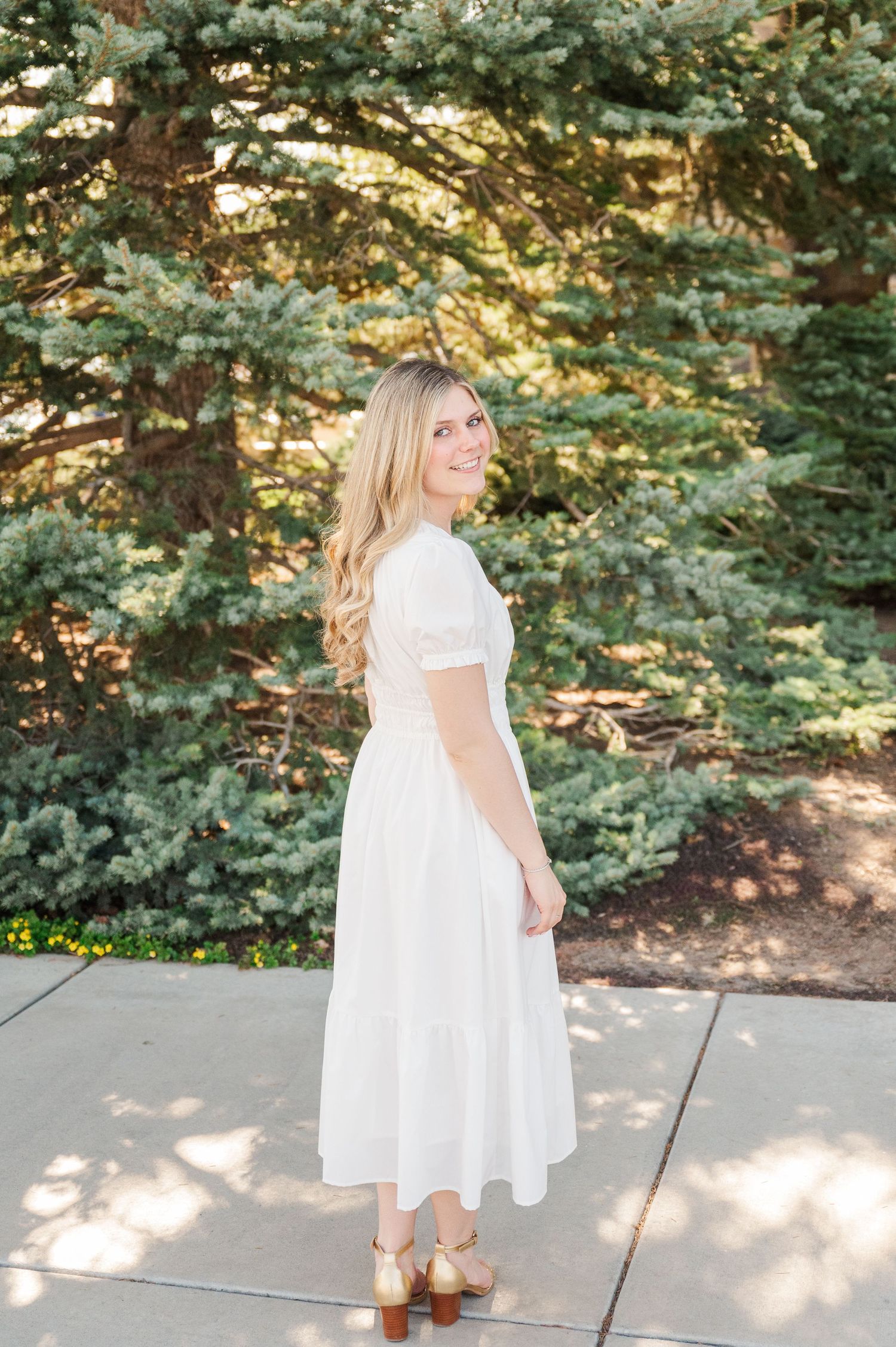 Woman in white dress poses outdoors against backdrop of evergreen trees.