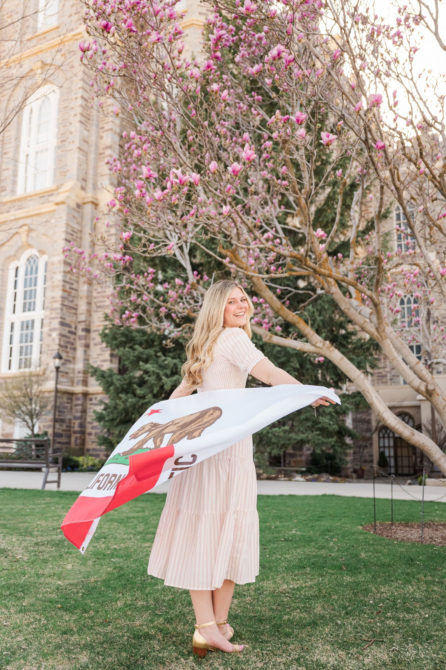 Woman twirls with California flag on lawn near blooming magnolia tree and stone building.