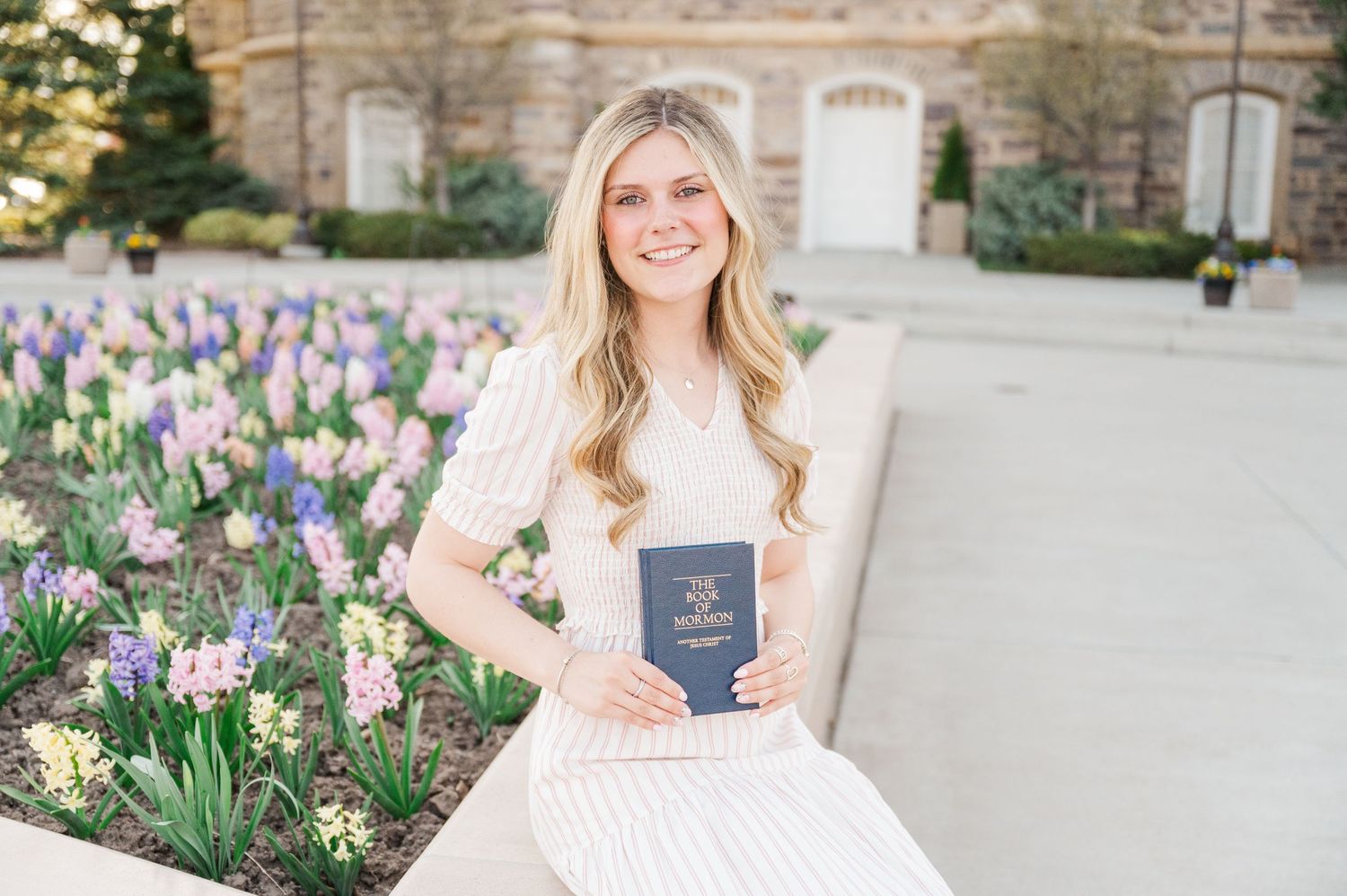 Woman holds book while standing near blooming spring tulips outside stone building.