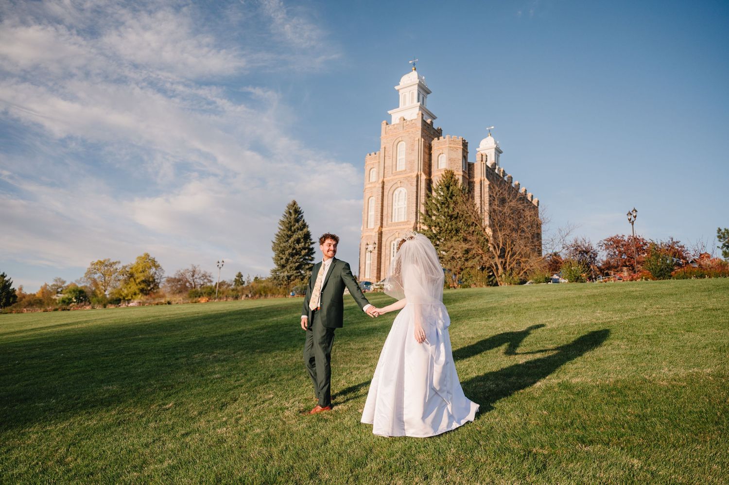 Two people in wedding attire holding hands while walking on green lawn in front of Logan Utah Temple on autumn day.