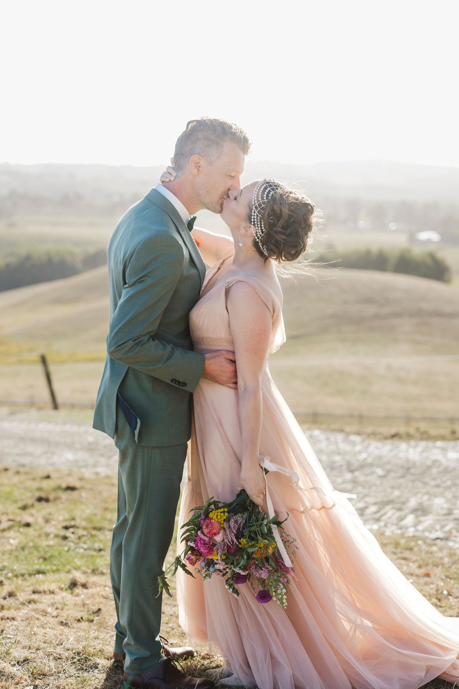 A couple embracing on a hilltop in formal attire with a blush pink wedding dress.