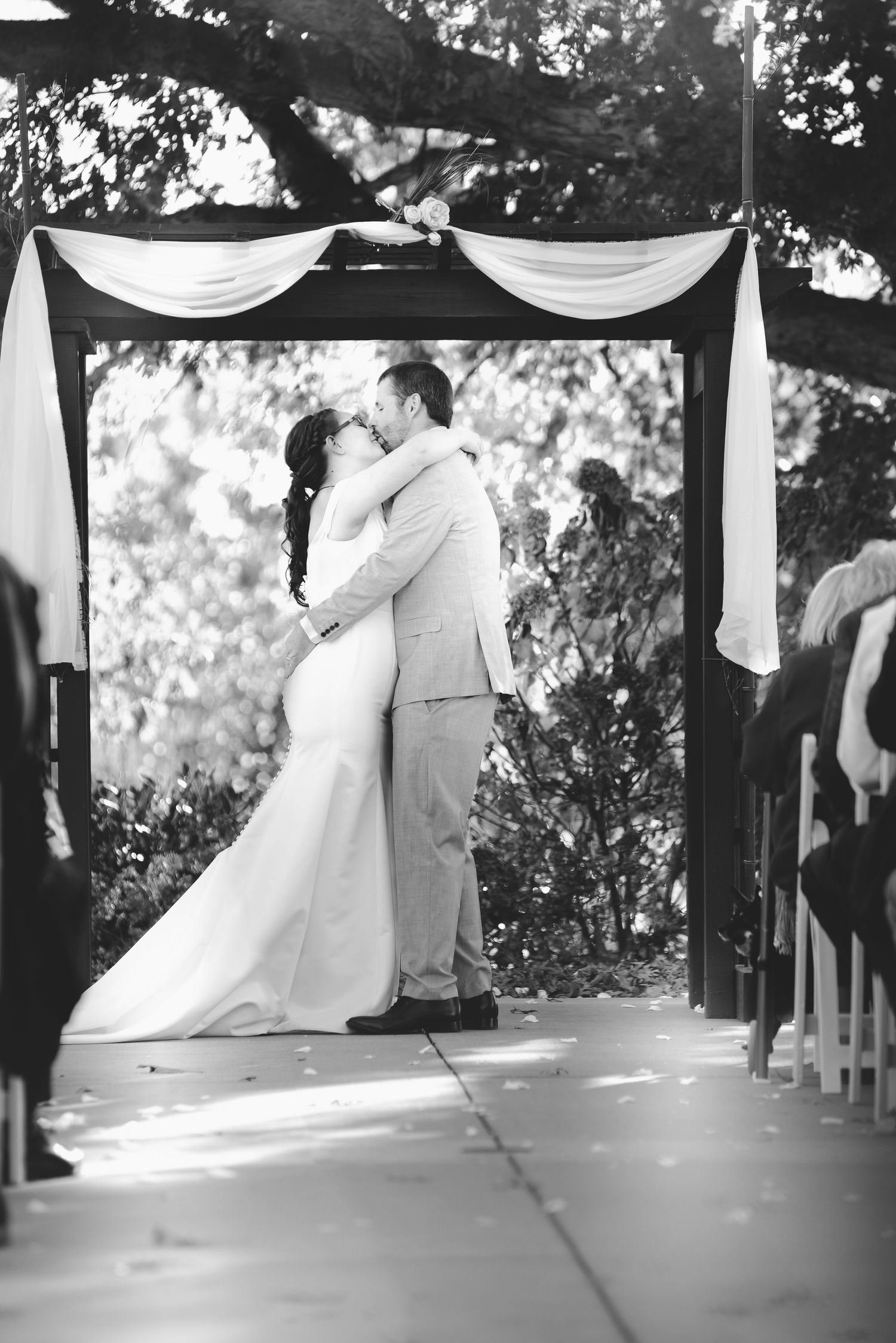 A romantic wedding ceremony takes place under a draped canopy arch in a beautiful outdoor garden setting.