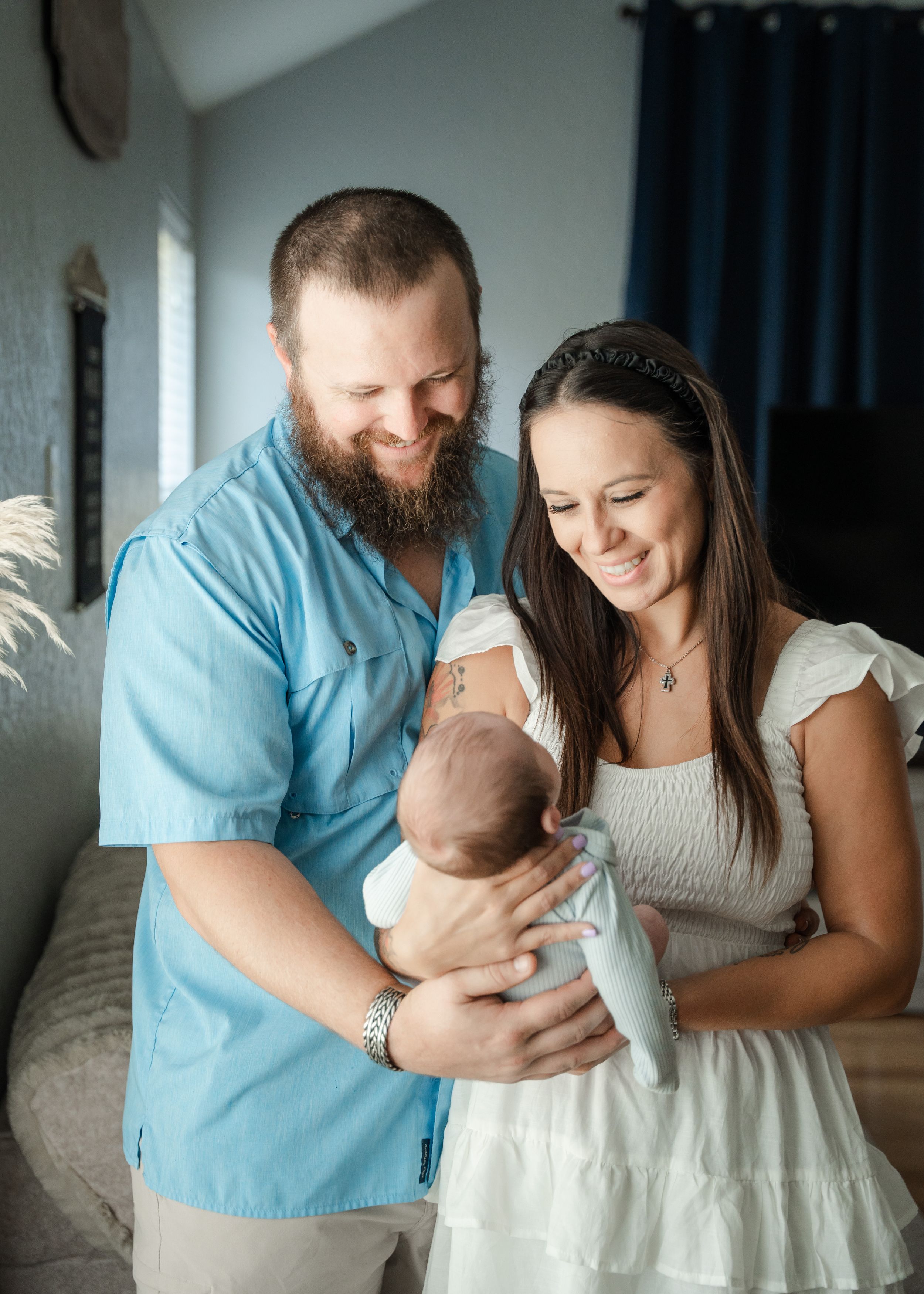 Happy couple with beards and white dress smiling at their newborn baby in a cozy home setting.