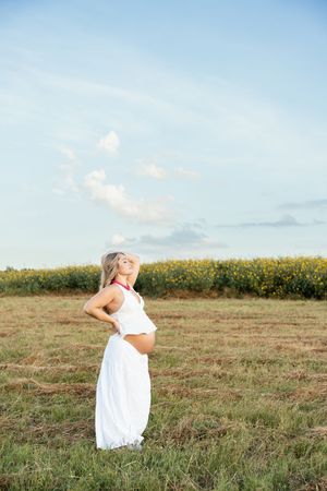 Pregnant blonde woman in white dress standing in open field with sunflowers under blue sky.