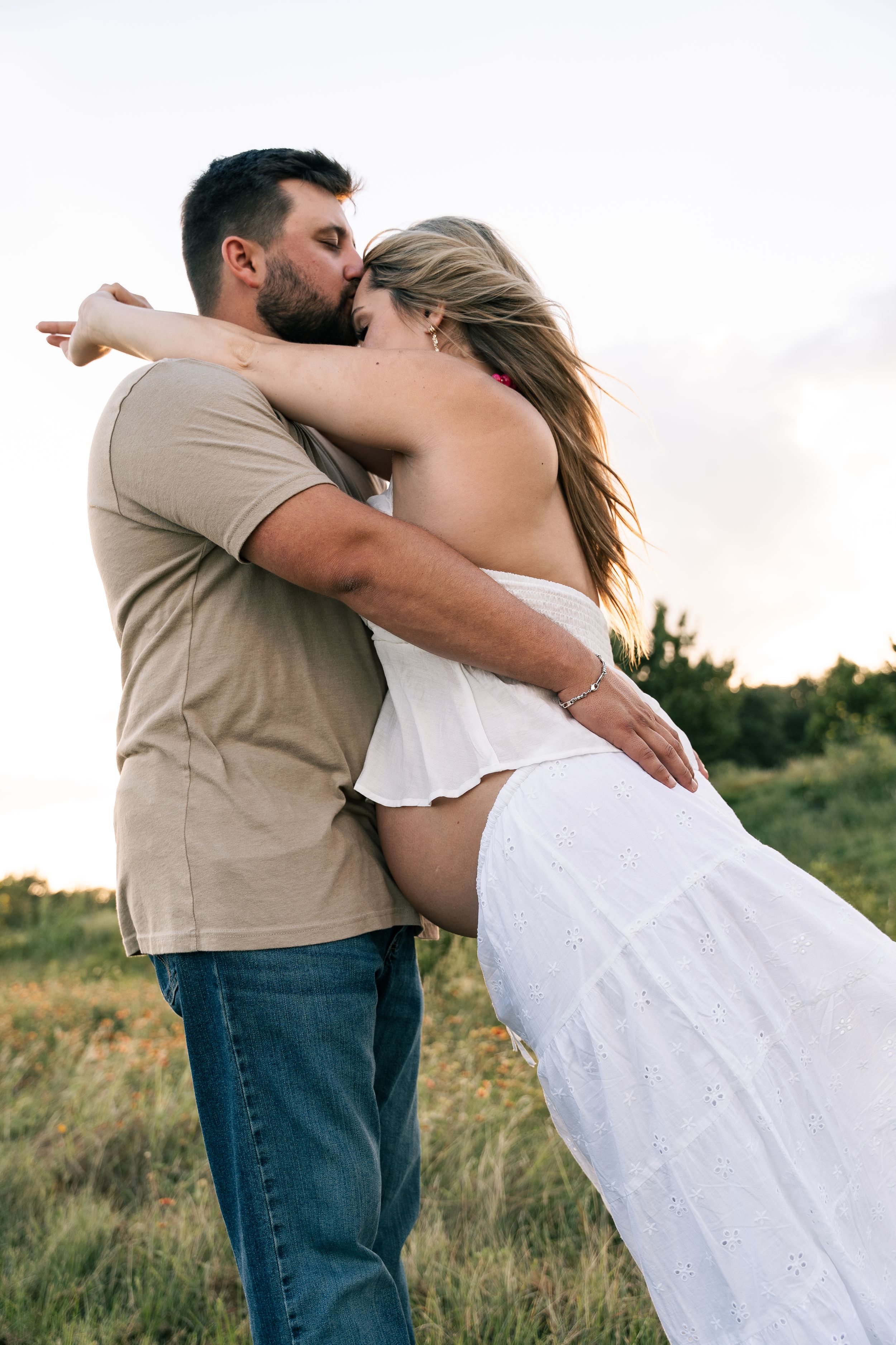 Couple sharing a romantic kiss in a golden field at sunset, woman in white dress, man in beige shirt and jeans.