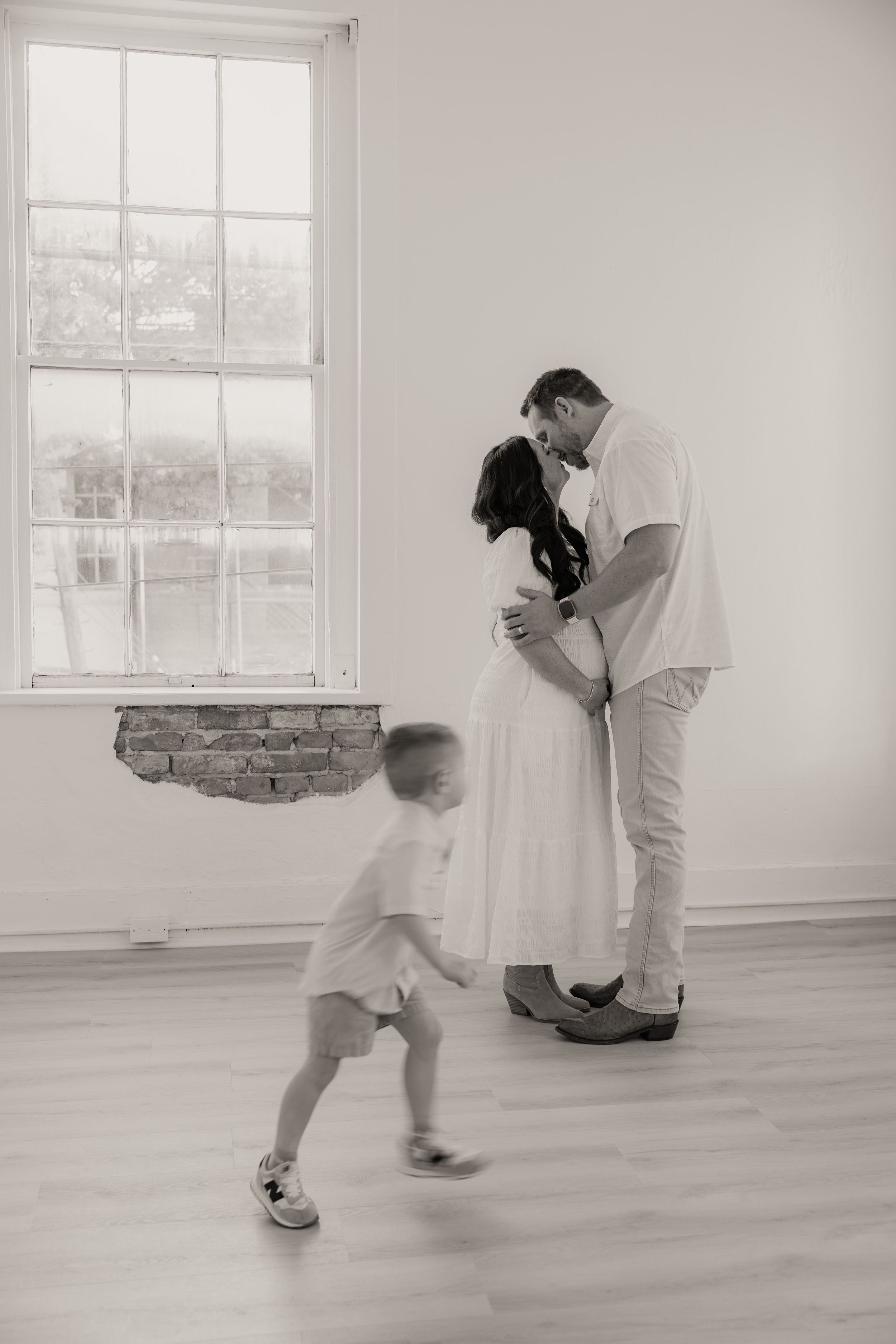 Black and white photo of pregnant couple kissing near window while toddler runs toward them.
