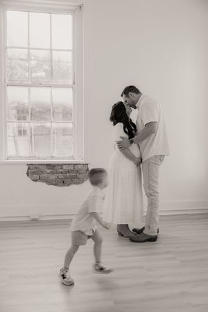 Black and white photo of pregnant couple kissing near window while toddler runs toward them.