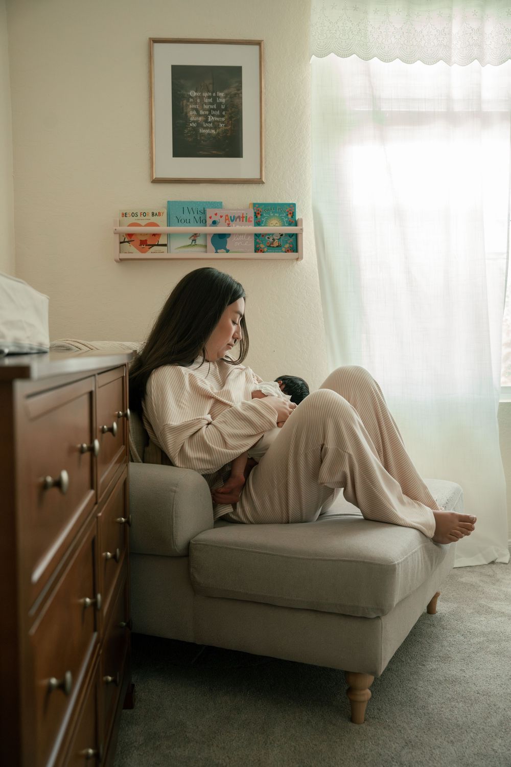 New mother sitting in nursery chair cradling newborn baby surrounded by soft natural light.