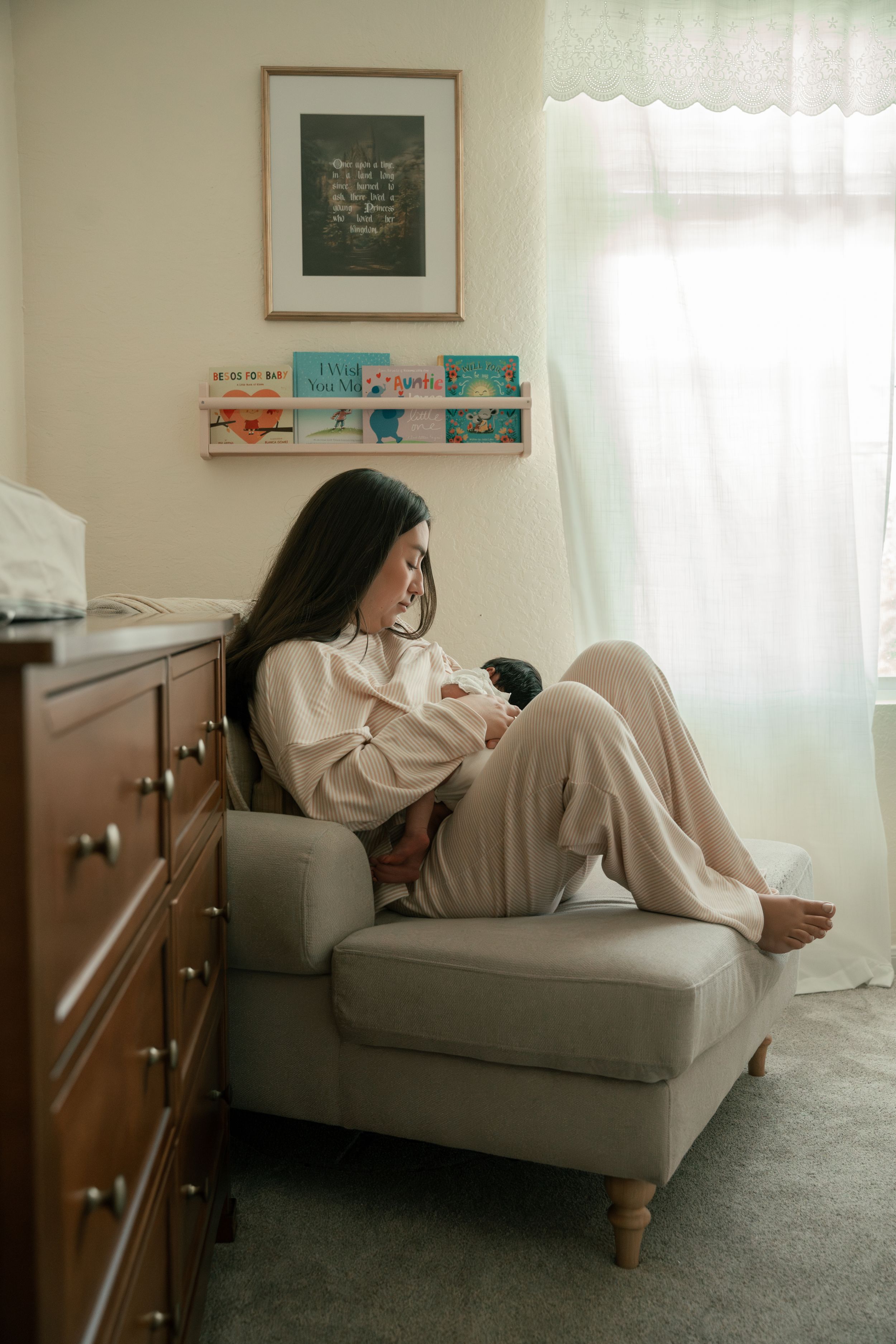 New mother sitting in nursery chair cradling newborn baby surrounded by soft natural light.