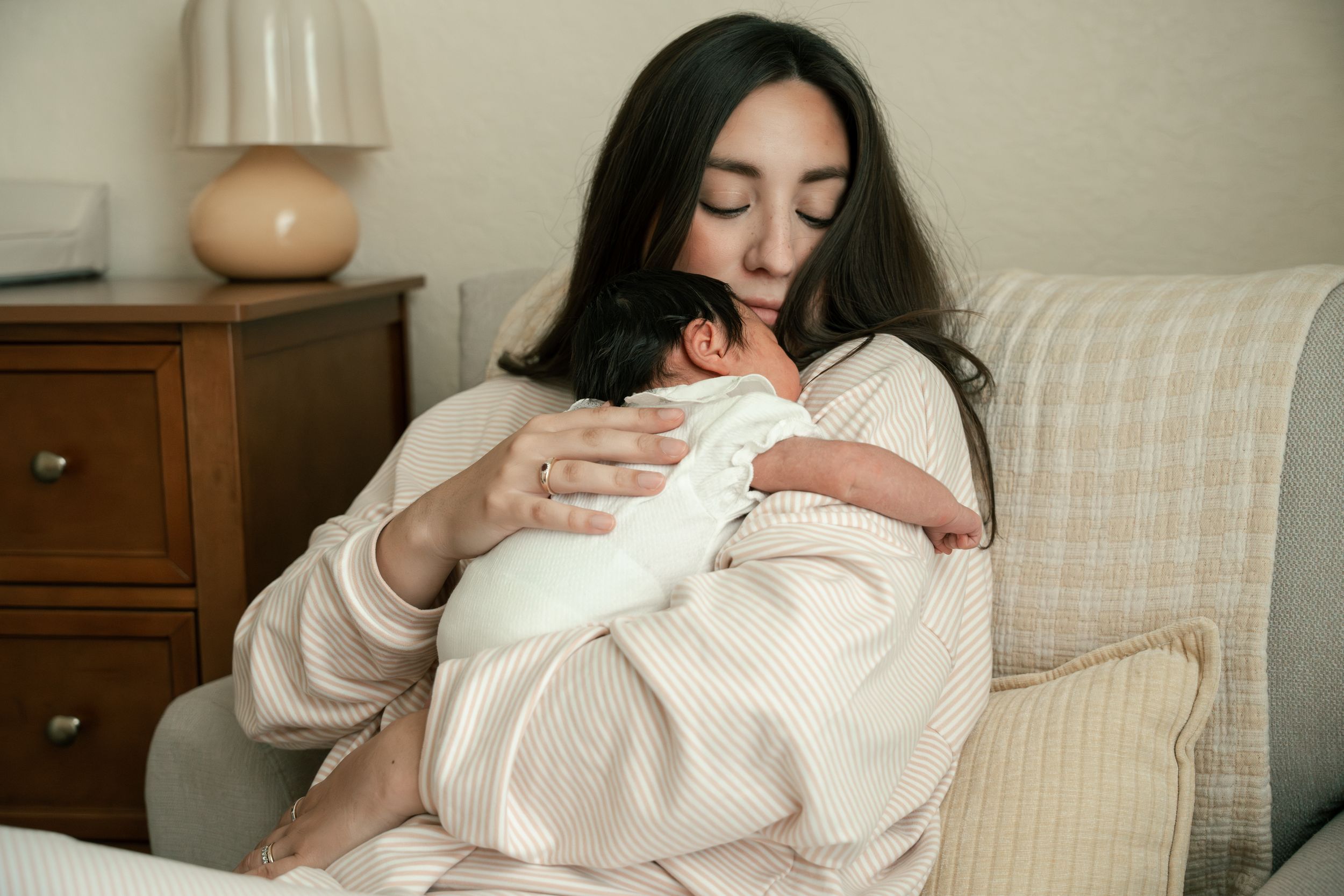 Dark-haired mother tenderly holding sleeping newborn baby dressed in white against her chest.