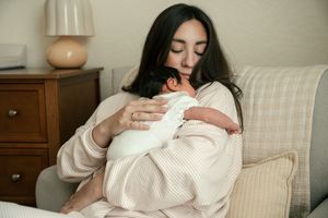 Dark-haired mother tenderly holding sleeping newborn baby dressed in white against her chest.