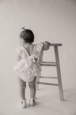 Black and white photo of a baby girl in a white ruffle romper standing at a wooden stool.