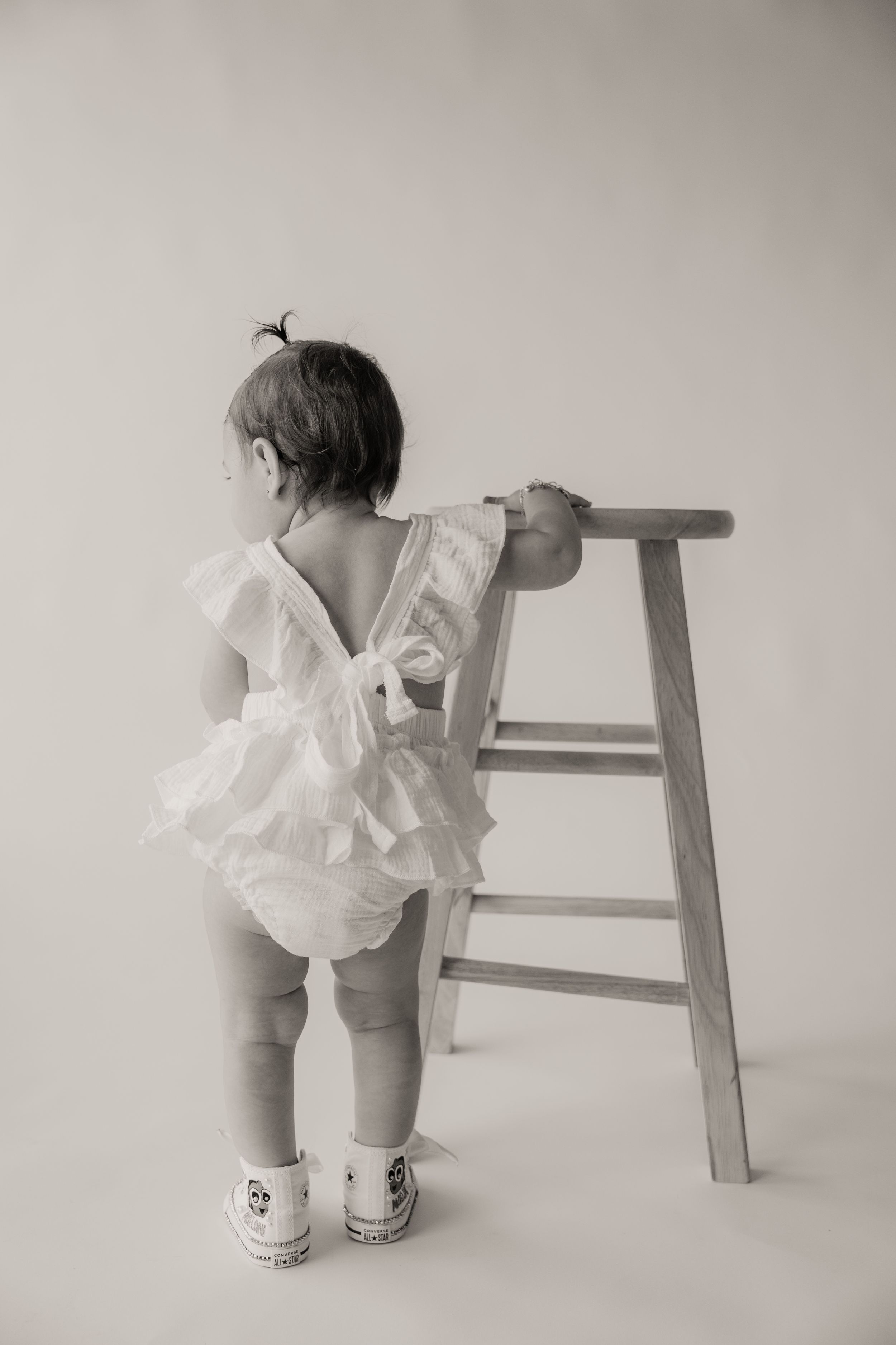 Black and white photo of a baby girl in a white ruffle romper standing at a wooden stool.