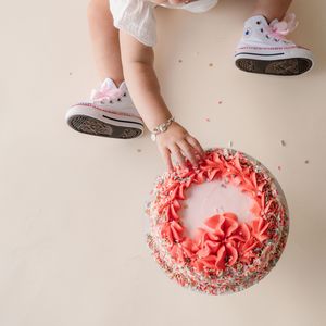 Overhead view of a baby's hands touching a pink floral birthday cake while wearing Converse sneakers.