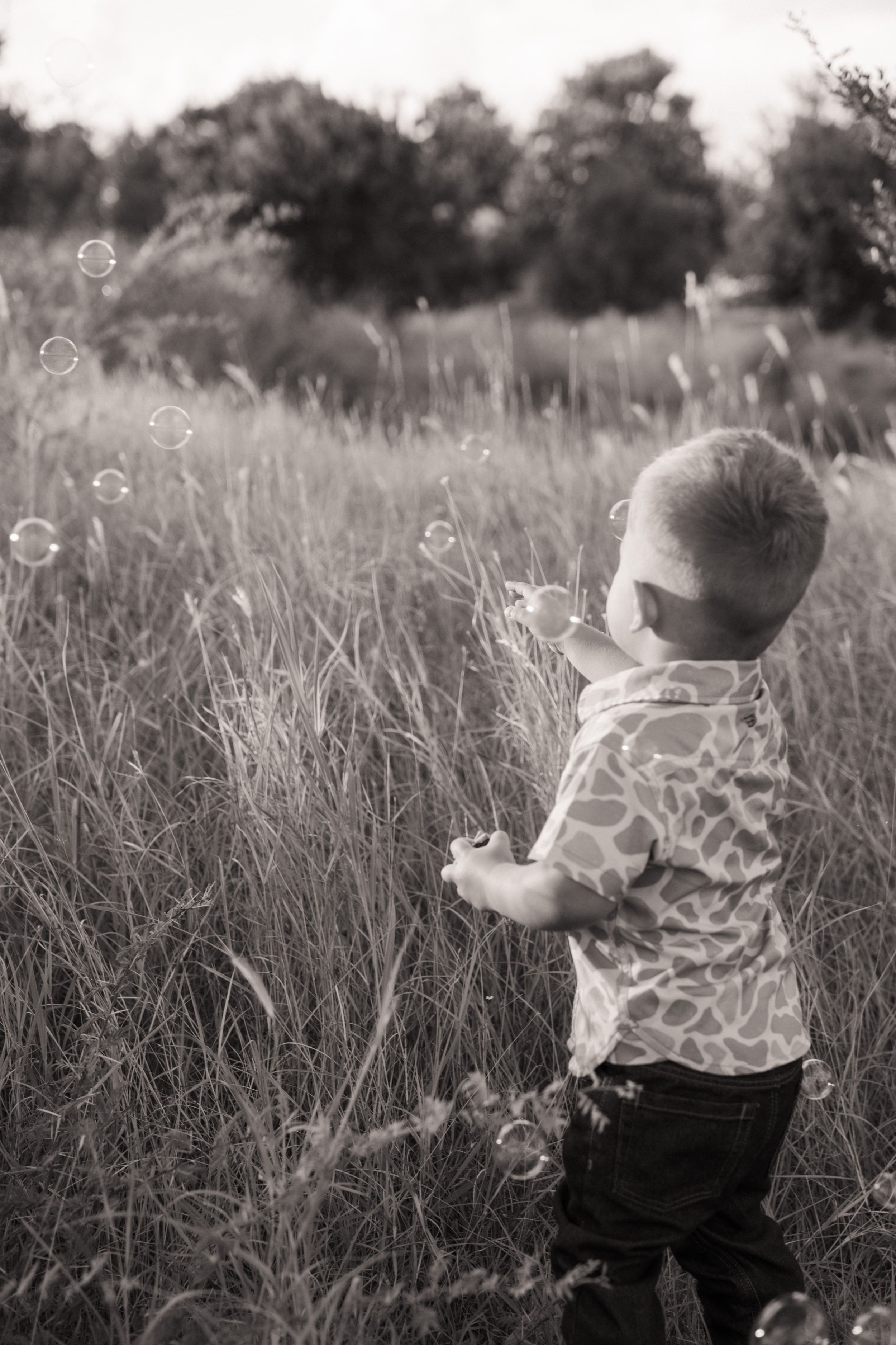 Black and white photo of a young boy playing with bubbles in a tall grass field outdoors.