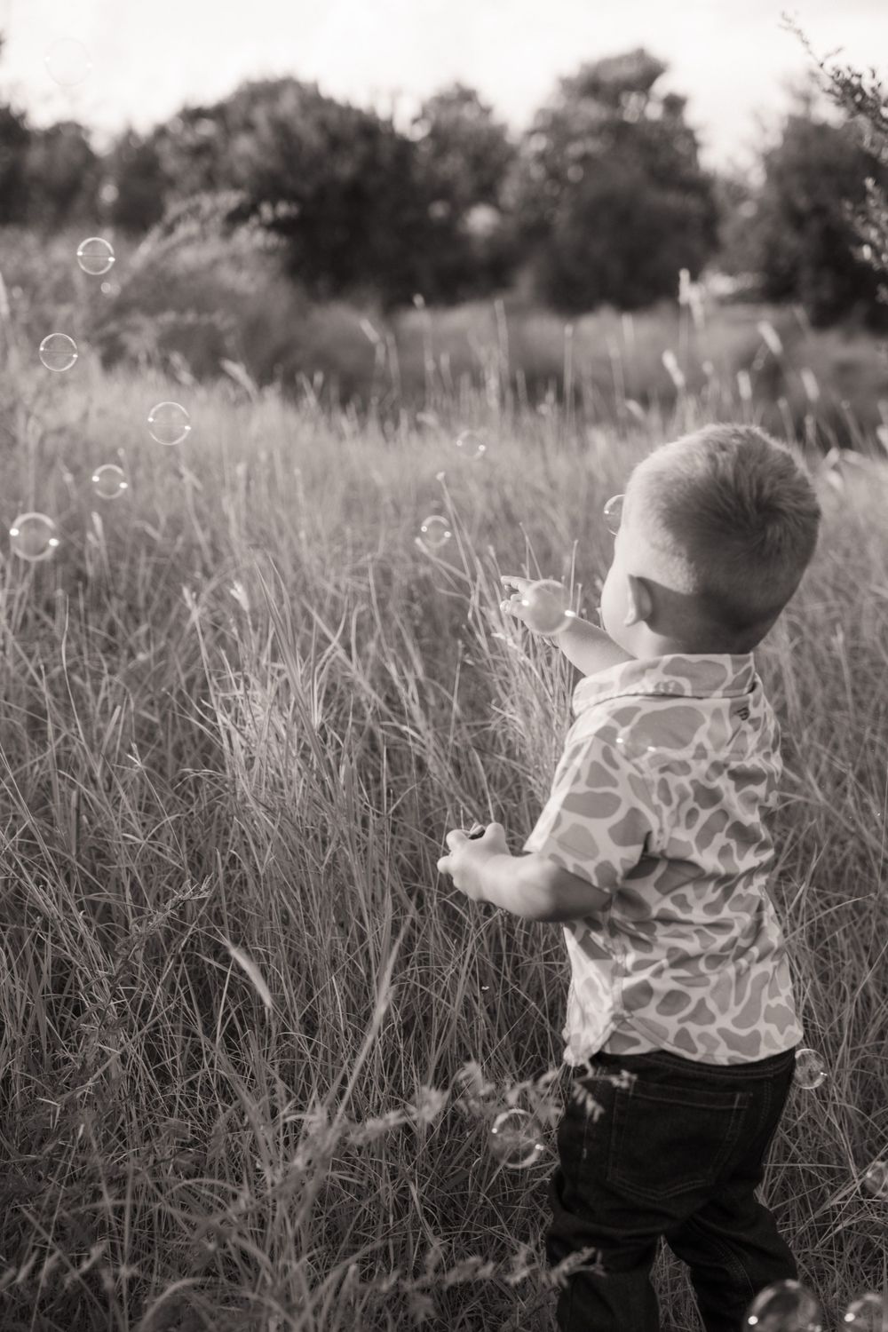 Black and white photo of a young boy playing with bubbles in a tall grass field outdoors.