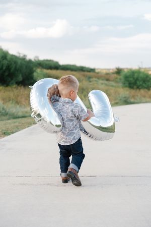 Young boy in giraffe print shirt carrying large silver balloon while walking on a path outdoors.