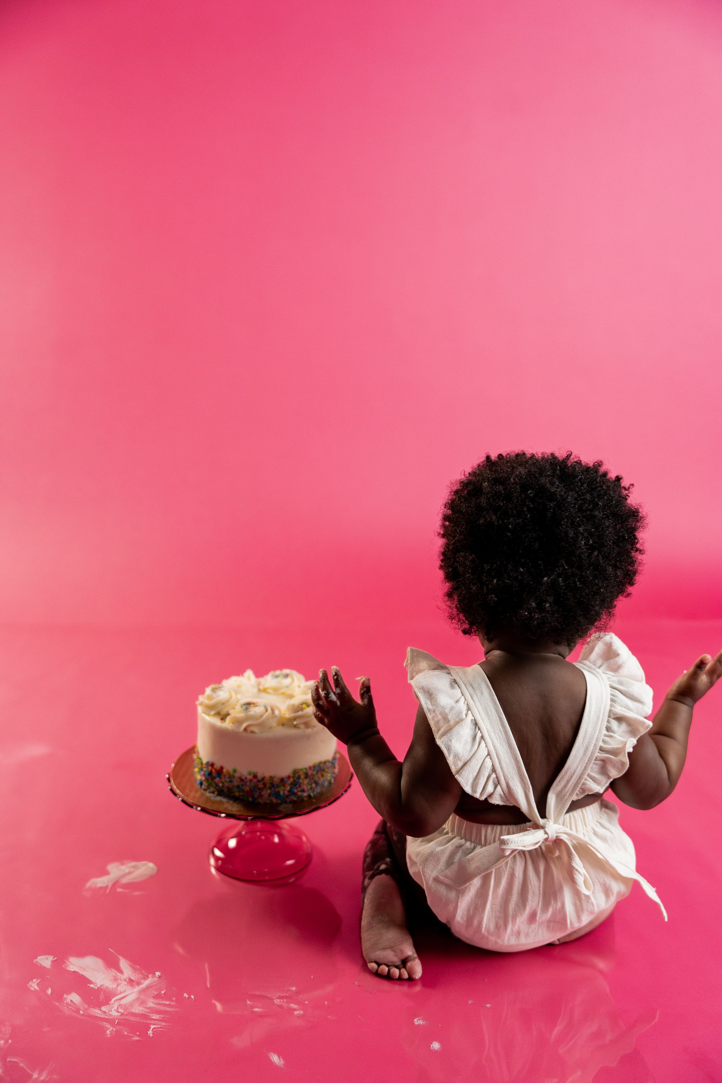 African American baby girl in white dress sitting before first birthday cake on pink background during 1st birthday milestone session.