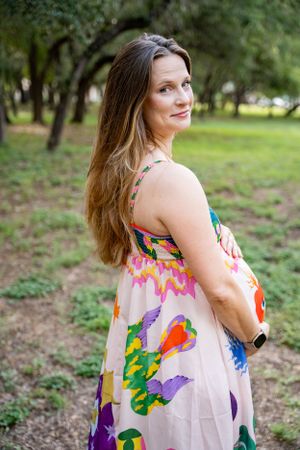 Pregnant woman in colorful floral dress smiling over shoulder in a lush green park.