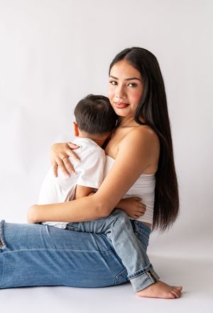 Beautiful mother sitting on white background hugging her young son, wearing white top and jeans.