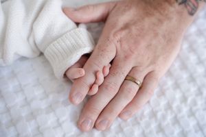 Close-up of newborn baby holding adult finger, with tattooed parent hand wearing gold wedding band.