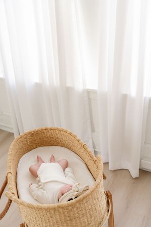 Newborn baby sleeping peacefully in a natural woven Moses basket bassinet near sheer white curtains.