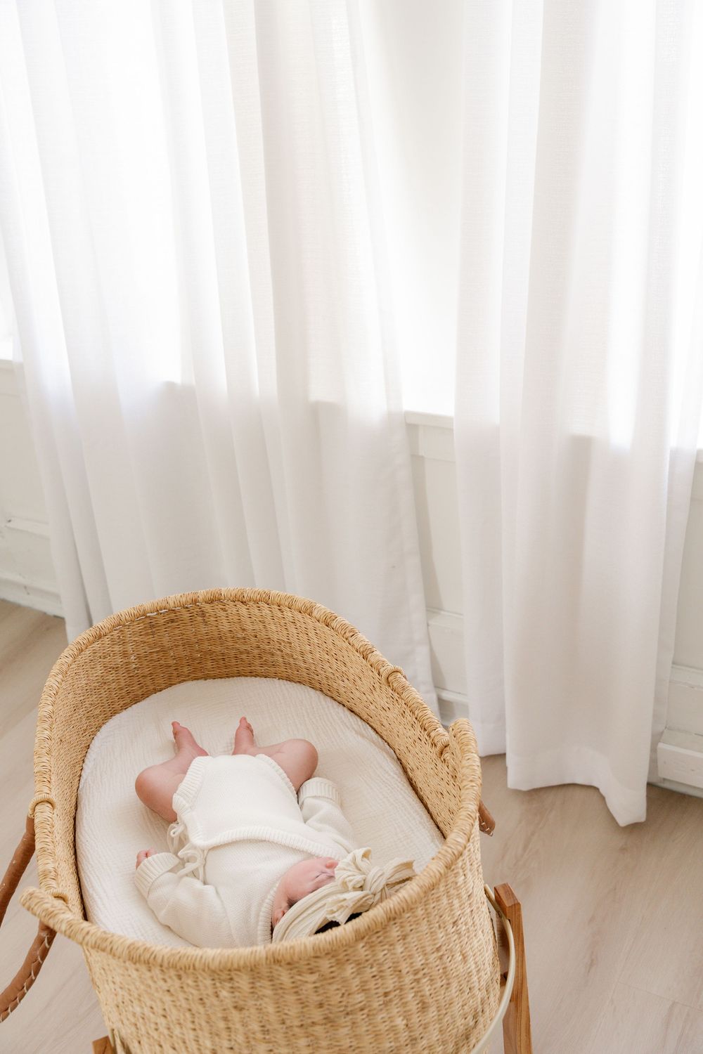 Newborn baby sleeping peacefully in a natural woven Moses basket bassinet near sheer white curtains.